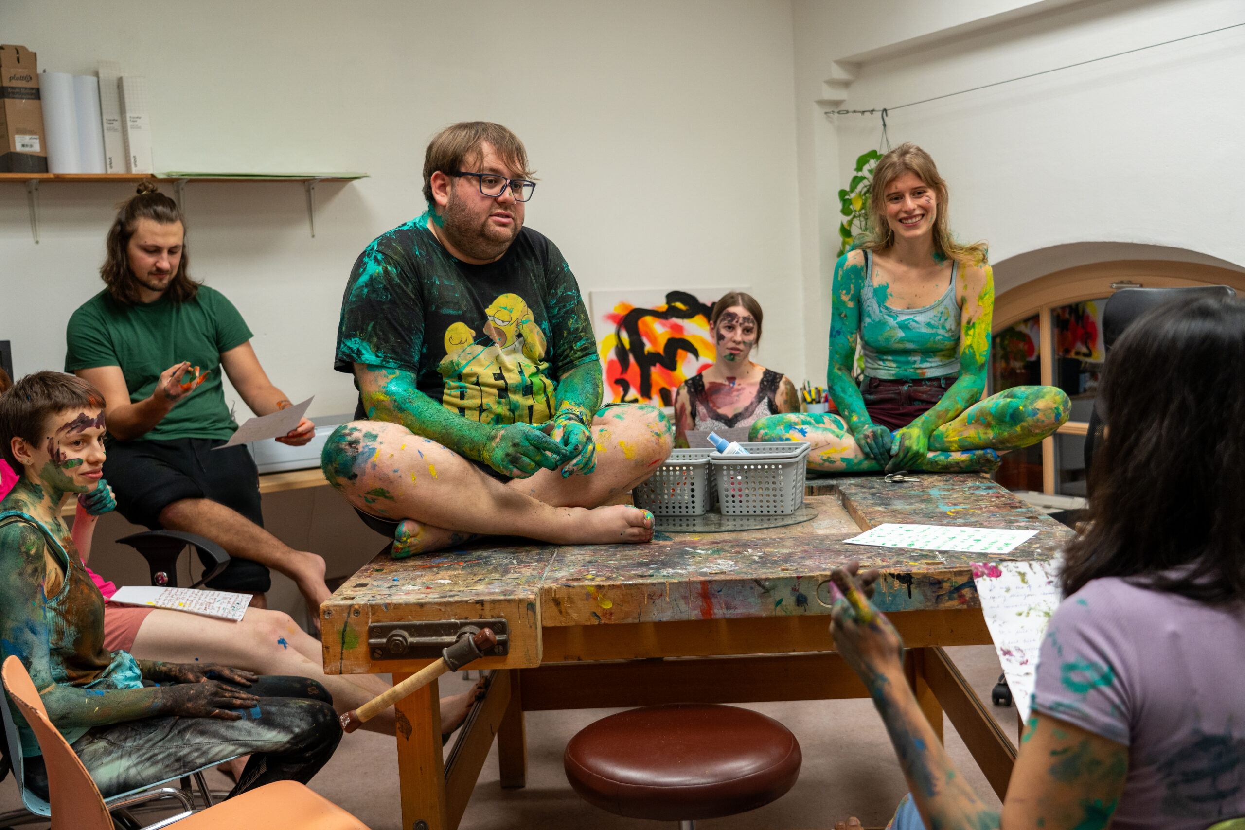 Eine Gruppe von fünf Personen, bedeckt mit bunter Farbe, sitzt um einen unordentlichen Tisch in einem Kunstatelier und unterhält sich. Umgeben von Kunstzubehör und bunten Kunstwerken erkunden sie Kunst und Angewandtes Theater, während die Kreativität in jede Ecke fließt. at Hochschule Coburg