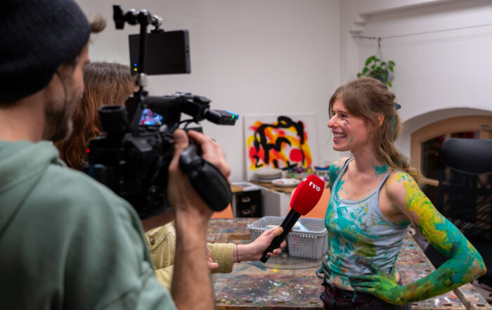 Eine lächelnde Frau mit Farbe an Armen und Kleidung wird im Fernsehen interviewt. Sie steht in einem bunten Kunststudio und hält ein rotes Mikrofon in der Hand, während zwei Personen sie filmen und aufnehmen. at Hochschule Coburg