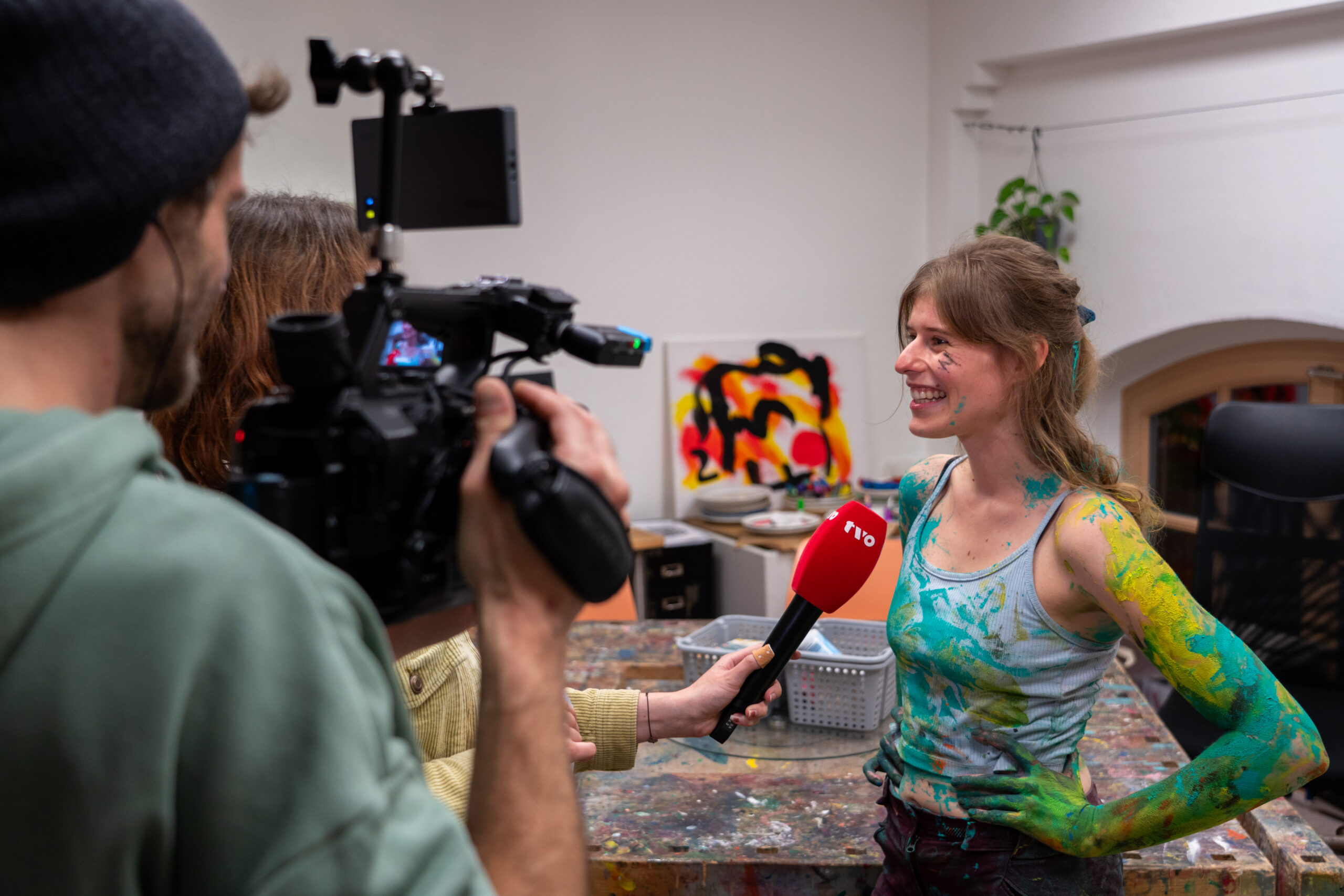 Eine lächelnde Frau mit Farbe an Armen und Kleidung wird im Fernsehen interviewt. Sie steht in einem bunten Kunststudio und hält ein rotes Mikrofon in der Hand, während zwei Personen sie filmen und aufnehmen. at Hochschule Coburg