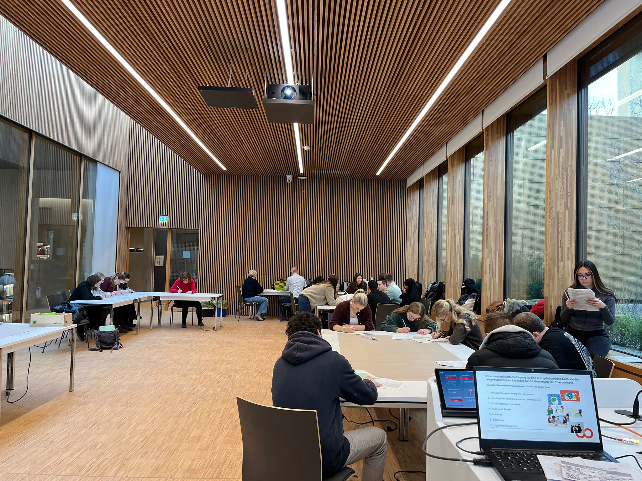 Eine Gruppe von Menschen sitzt an Tischen in einem modernen, holzgetäfelten Raum und nimmt an einem Workshop teil, der wissenschaftliche Klimafakten spielerisch vermittelt. Ein Laptop mit einer bunten Präsentation steht im Vordergrund. Große Fenster lassen natürliches Licht herein. at Hochschule Coburg