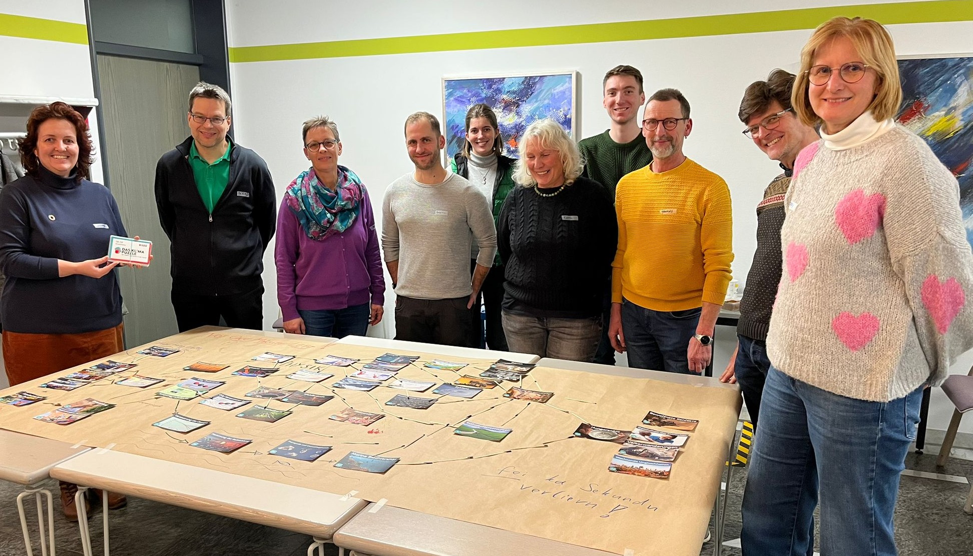 Zehn Erwachsene stehen um einen mit Karten und Bildern bedeckten Tisch in einem Workshop und lächeln in die Kamera. In diesem hell erleuchteten Raum mit bunten Kunstwerken werden wissenschaftliche Klimafakten spielerisch vermittelt. at Hochschule Coburg