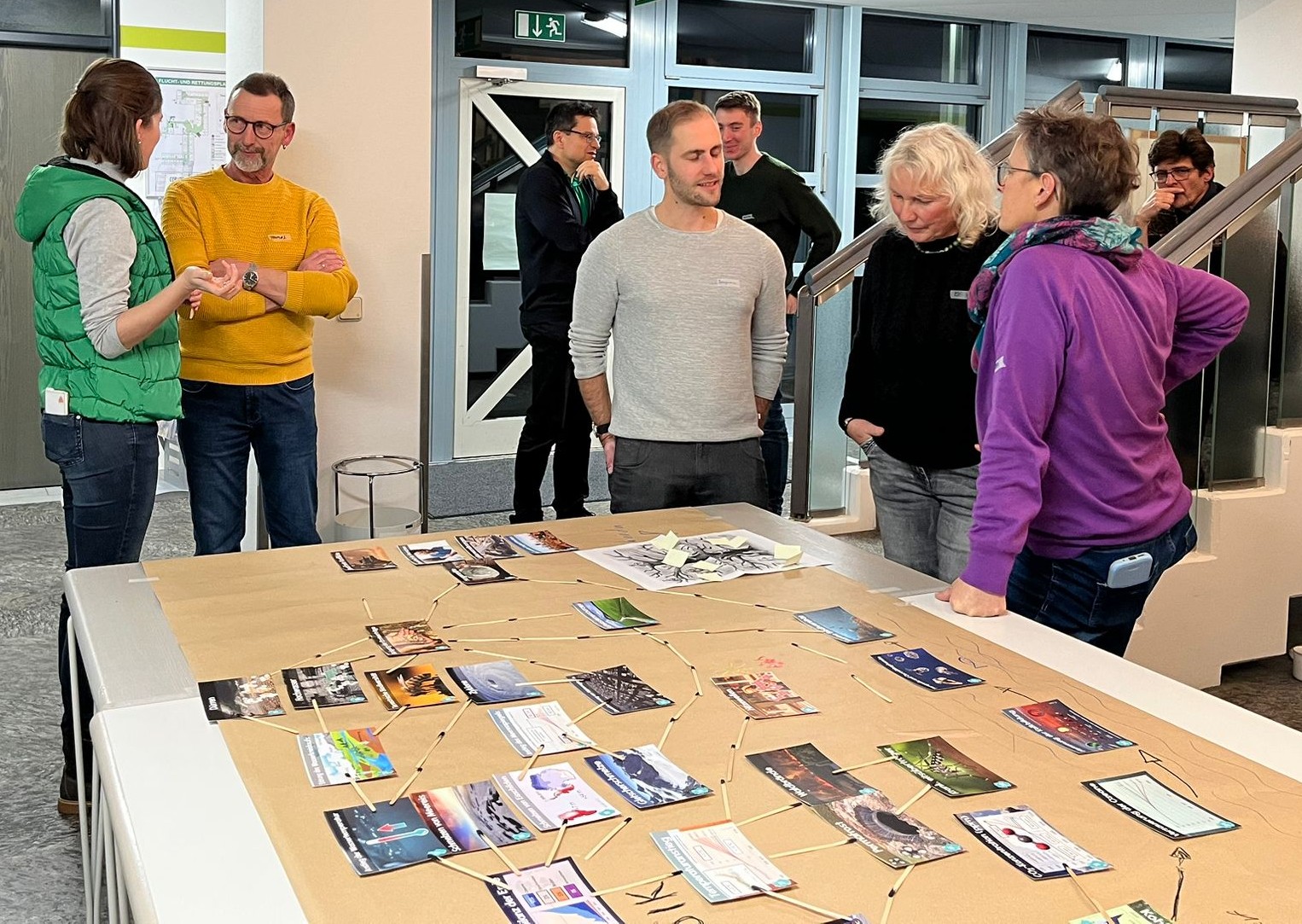 Eine Gruppe von Menschen diskutiert und betrachtet einen mit Fotos und Diagrammen bedeckten Tisch in einem hell erleuchteten Innenraum, was auf einen gemeinsamen Workshop zur spielerisch wissenschaftlichen Klimafaktenvermittlung hindeutet. at Hochschule Coburg