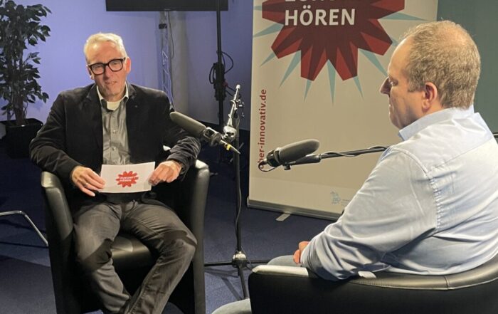 Zwei Männer sitzen sich auf schwarzen Stühlen gegenüber und nehmen in einem Studio mit Mikrofonen einen Podcast auf. Hinter ihnen ist ein Transparent mit der Aufschrift "Zukunft Hören" zu sehen. Studiolampen und Aufnahmegeräte, darunter ein automatisch gespeicherter Entwurf, sind zu sehen. at Hochschule Coburg