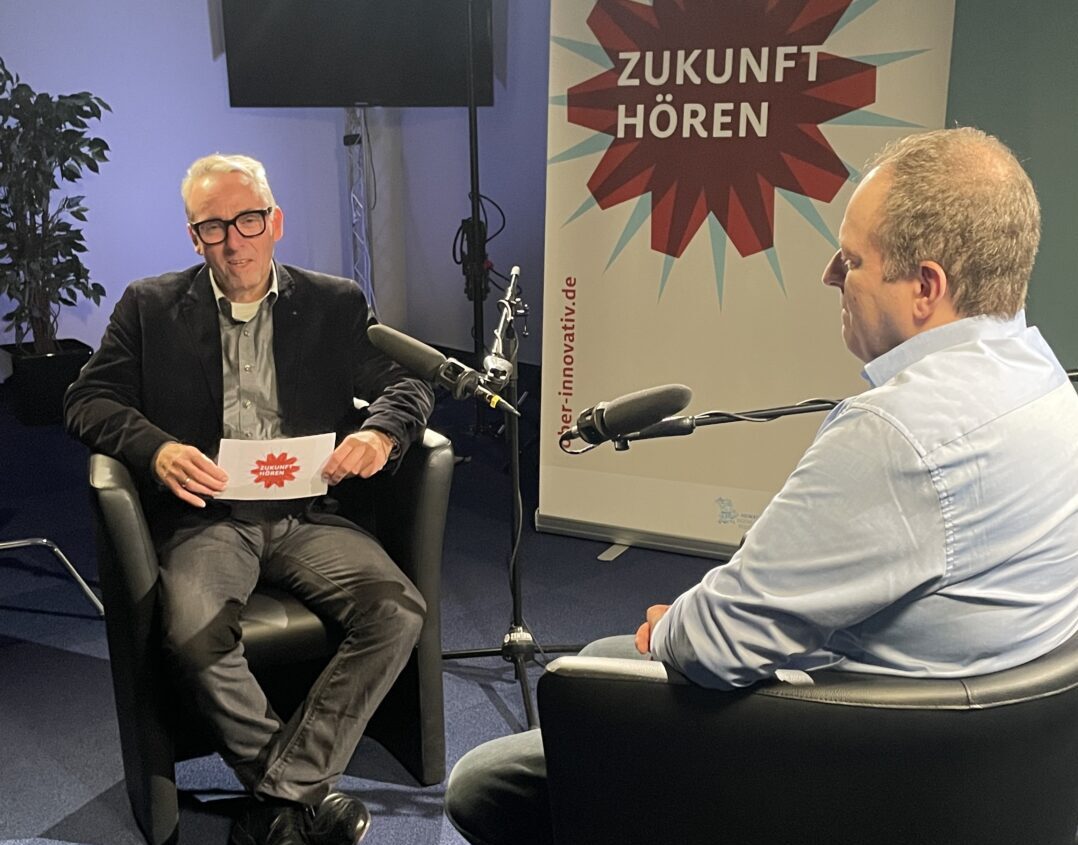 Zwei Männer sitzen sich auf schwarzen Stühlen gegenüber und nehmen in einem Studio mit Mikrofonen einen Podcast auf. Hinter ihnen ist ein Transparent mit der Aufschrift "Zukunft Hören" zu sehen. Studiolampen und Aufnahmegeräte, darunter ein automatisch gespeicherter Entwurf, sind zu sehen. at Hochschule Coburg