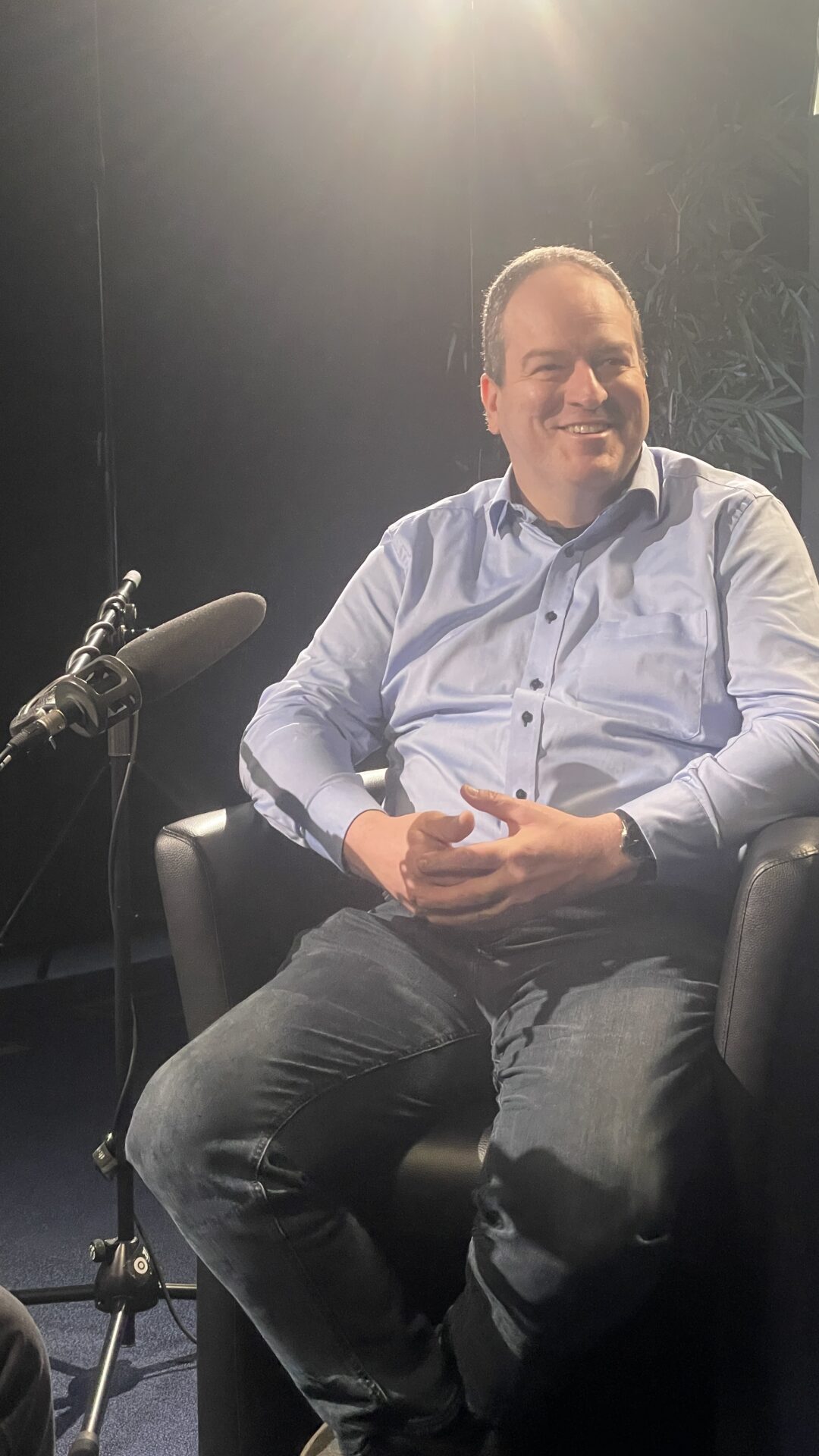 Ein Mann in hellblauem Hemd und Jeans sitzt lächelnd in einem schwarzen Sessel und hat ein Mikrofon vor sich stehen. Der Hintergrund ist schwach beleuchtet, mit einer hellen Lichtquelle hinter ihm. at Hochschule Coburg