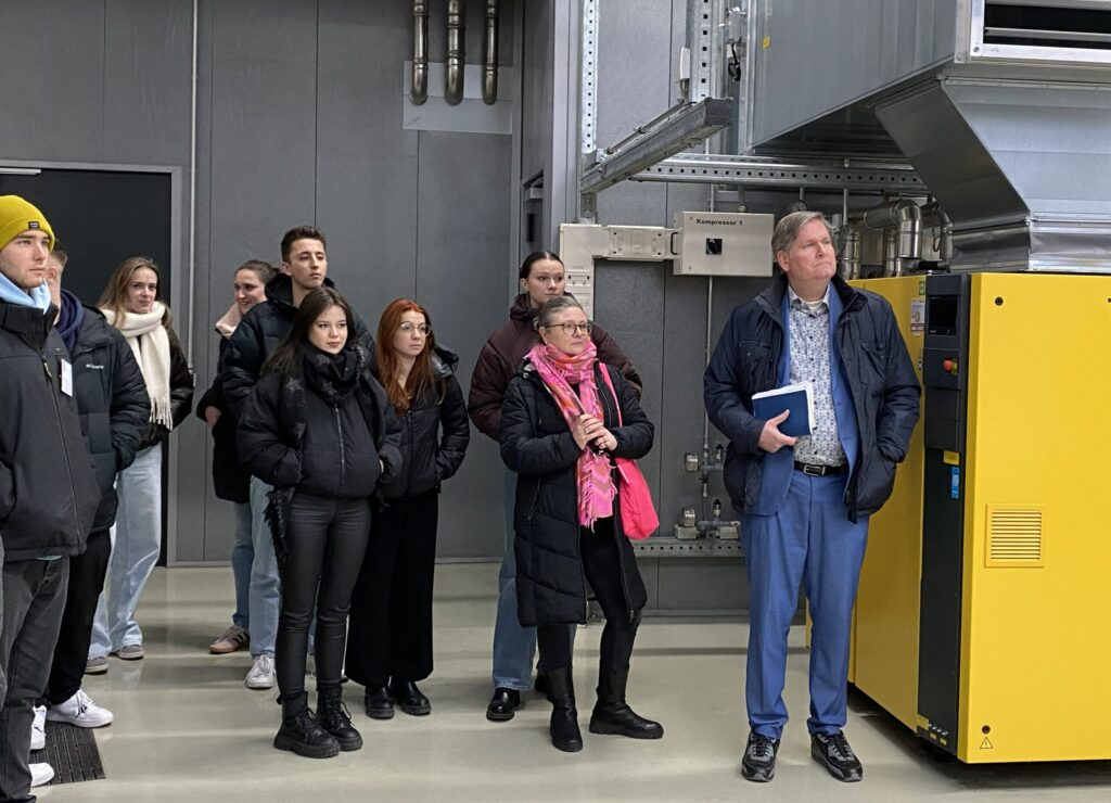 Eine Gruppe von Personen, darunter ein Mann im blauen Anzug, der einen Ordner in der Hand hält, steht in einem Innenraum in der Nähe von Industrieanlagen und beobachtet aufmerksam etwas aus dem Off - vielleicht eine Diskussion über Versicherungsmanagement oder Aspekte der Betriebswirtschaft. Die meisten sind in Winterkleidung gekleidet. at Hochschule Coburg