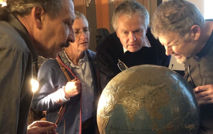 Vier Personen nehmen einen 200 Jahre alten Globus genau unter die Lupe und enthüllen auf einem mit blauem Tuch drapierten Tisch inmitten einer kunstvollen, warm beleuchteten Einrichtung Geschichte und Geheimnisse der Welt. at Hochschule Coburg