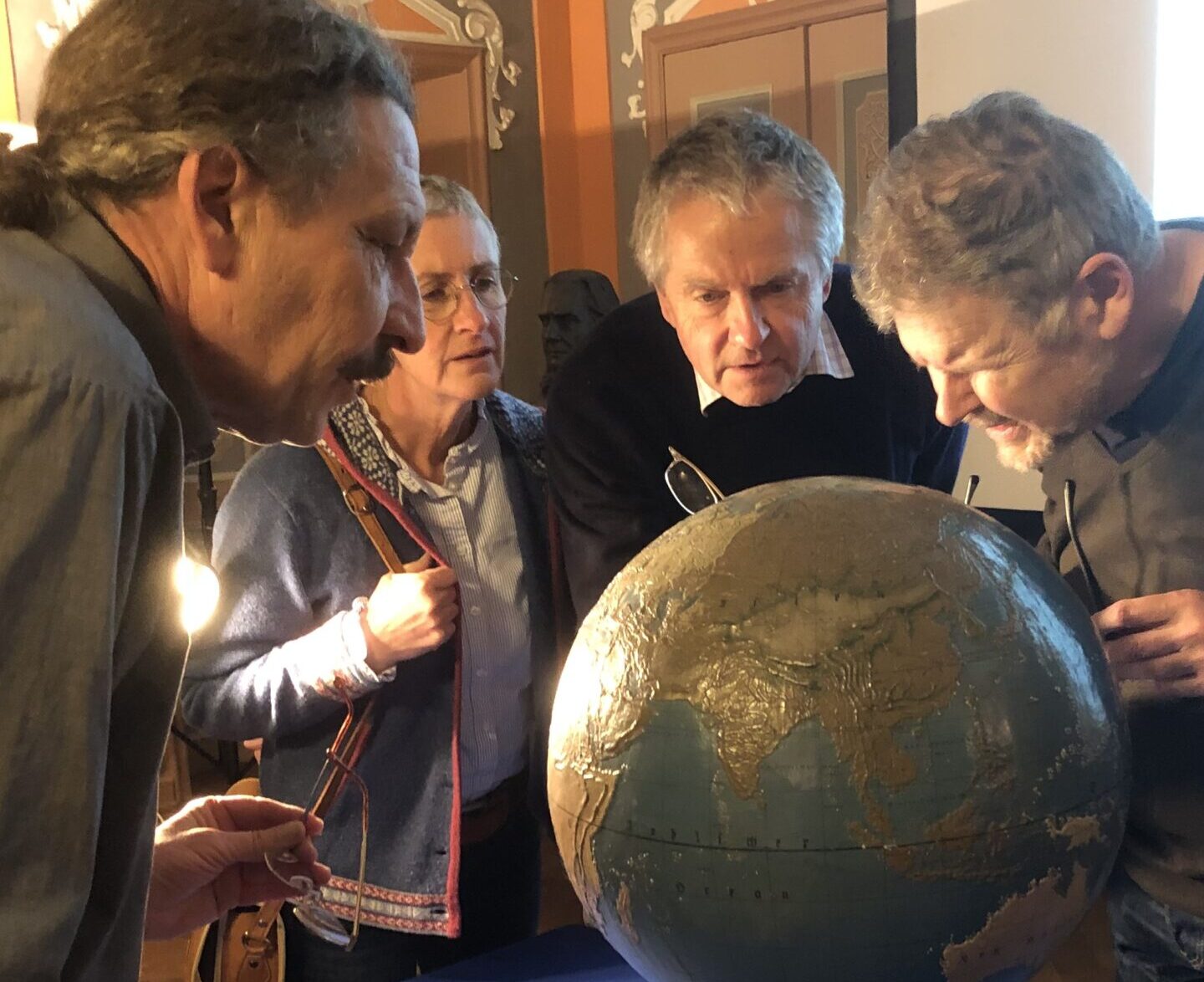Vier Personen nehmen einen 200 Jahre alten Globus genau unter die Lupe und enthüllen auf einem mit blauem Tuch drapierten Tisch inmitten einer kunstvollen, warm beleuchteten Einrichtung Geschichte und Geheimnisse der Welt. at Hochschule Coburg