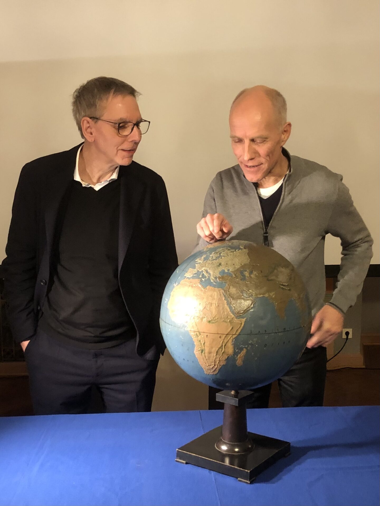 Zwei Männer stehen neben einem blauen Tisch und untersuchen einen alten Globus. Der eine zeigt auf den Globus, der andere beobachtet ihn, beide mit Brille und Pullover. Der sanft beleuchtete Hintergrund unterstreicht ihr Interesse an der Weltgeschichte. at Hochschule Coburg