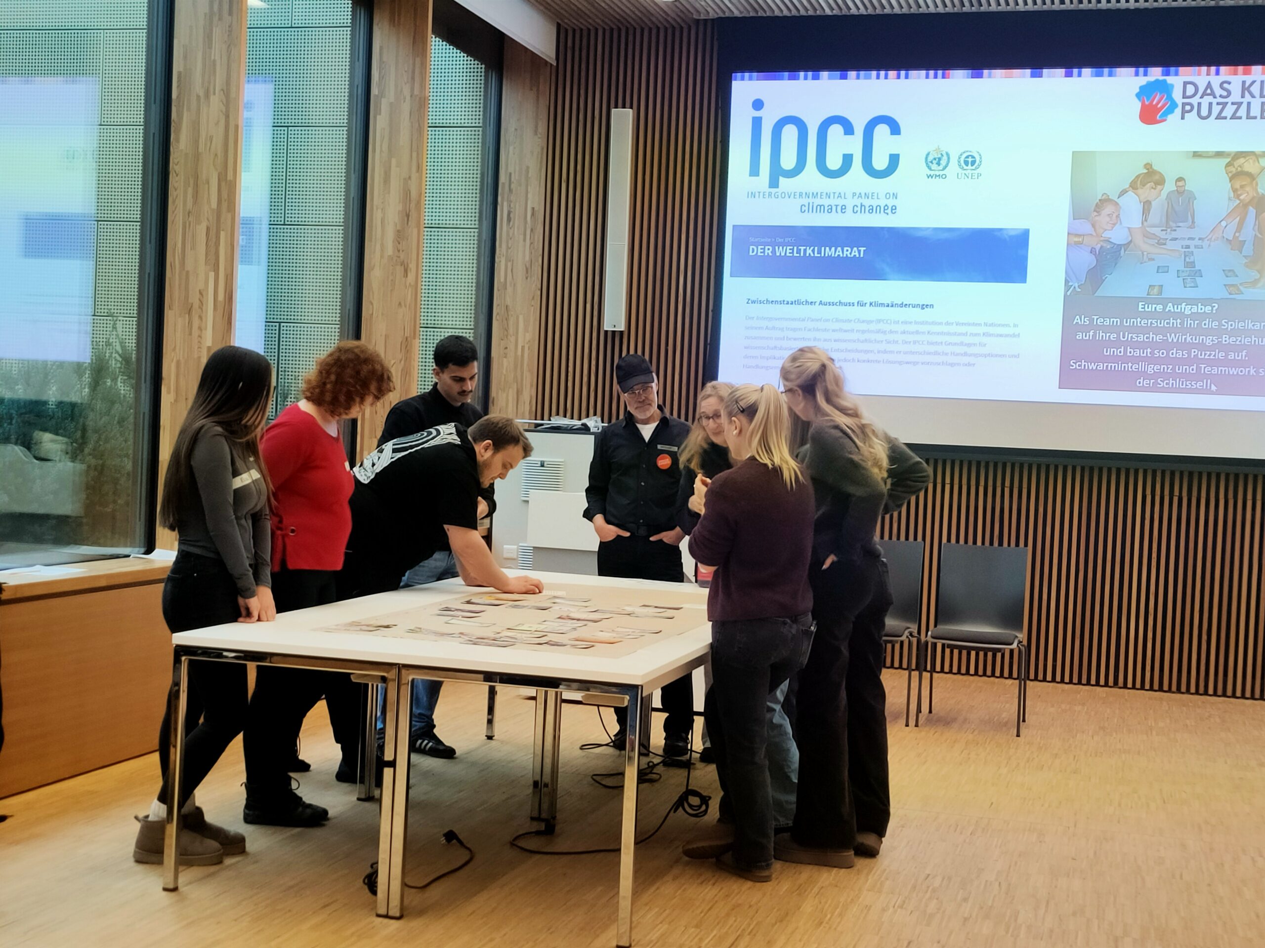 Sieben Personen stehen um einen Tisch herum und ordnen Karten, während hinter ihnen in einem modernen Konferenzraum wissenschaftliche Klimafakten spielerisch vermittelt werden, die auf Bildschirmen über den Klimawandel gezeigt werden. at Hochschule Coburg