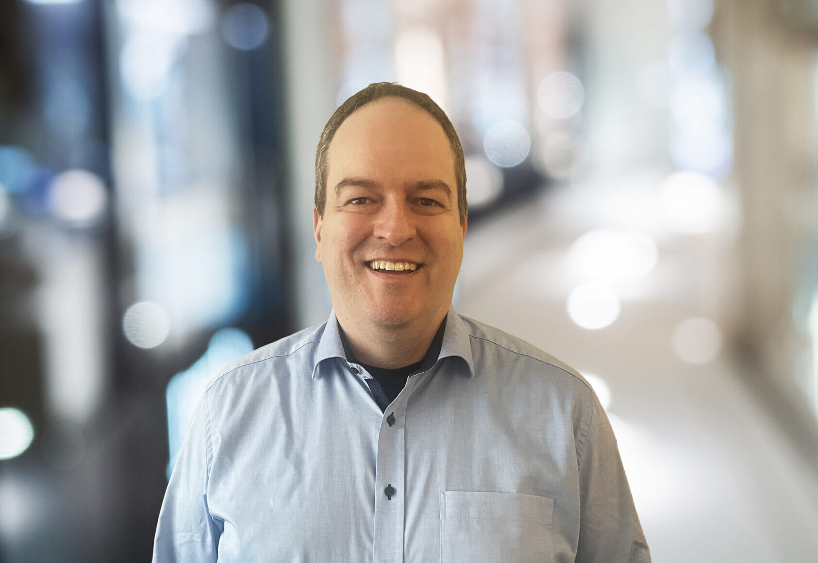 Ein Mann mit kurzen braunen Haaren, der ein hellblaues Hemd trägt, lächelt, während er in einem Innenraum steht. Der Hintergrund ist unscharf, wie bei einem automatisch gespeicherten Entwurf eines hellen, modernen Flurs mit Lichtern und Glasscheiben. at Hochschule Coburg