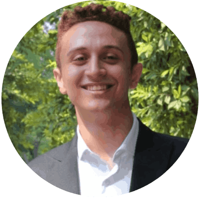 Ein junger Mann mit kurzem, lockigem Haar lächelt in die Kamera. Er trägt ein weißes Hemd und einen dunklen Blazer und steht im Freien mit grünem Laub im Hintergrund. at Hochschule Coburg