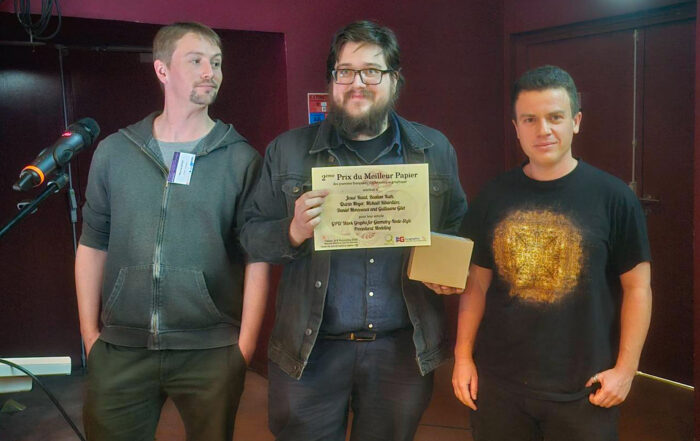 Drei Personen stehen in einem Raum neben einem Mikrofon. Die Person in der Mitte hält eine Urkunde und eine Schachtel in der Hand und lächelt, als würde sie einen automatisch gespeicherten Entwurf feiern. Die anderen stehen zu beiden Seiten, einer im Kapuzenpulli und einer im schwarzen T-Shirt mit goldenem Muster. at Hochschule Coburg