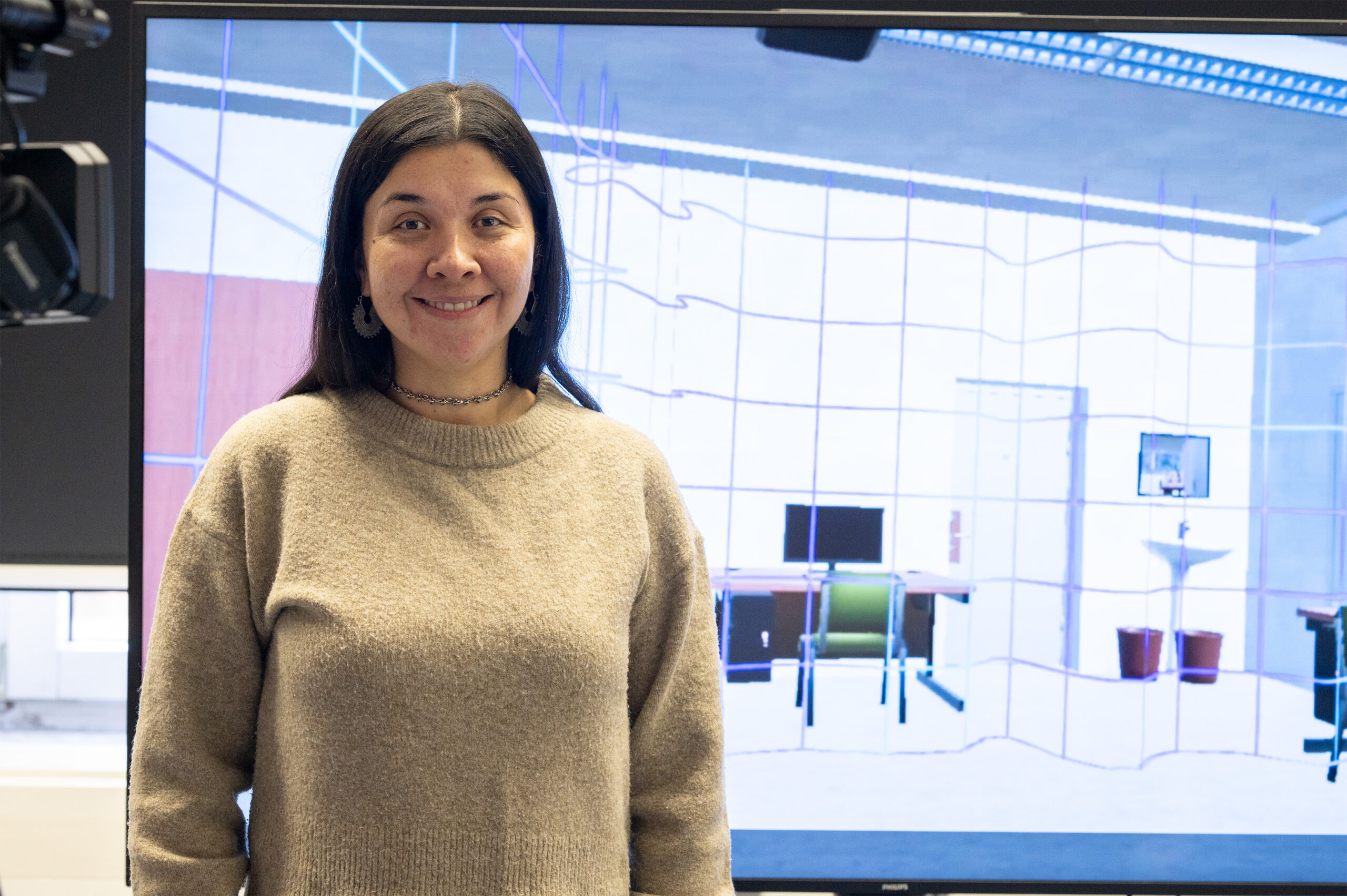 Eine Frau mit langen dunklen Haaren und einem beigen Pullover lächelt, während sie vor einem großen Bildschirm steht, auf dem ein digitales Büro- oder Arbeitsraumdesign zu sehen ist. at Hochschule Coburg