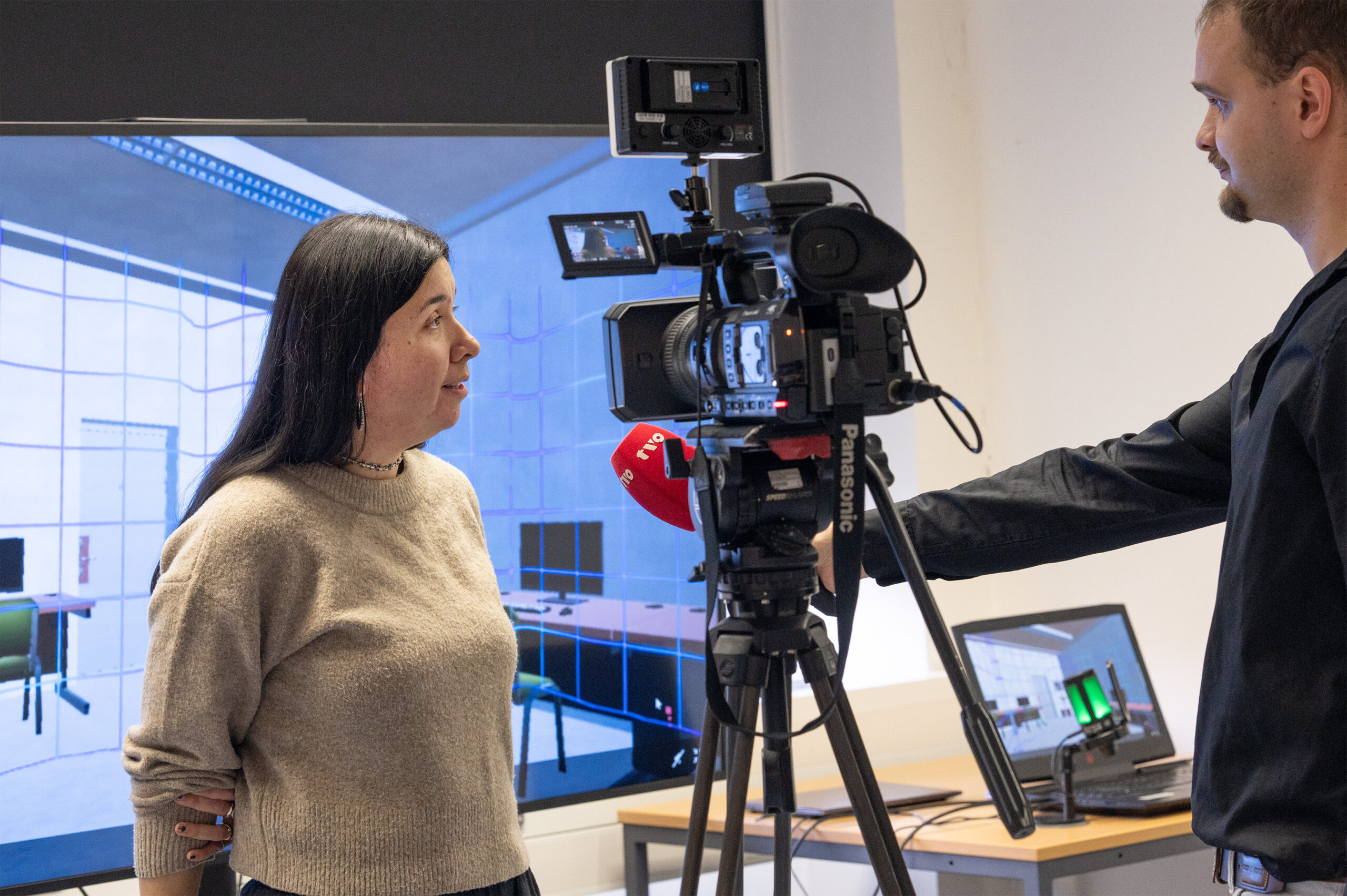 Eine Frau steht vor einer Videokamera und wird von einem Mann in einem schwarzen Hemd interviewt. Hinter ihnen ist ein großer Bildschirm zu sehen, der einen digitalen Raum mit Schreibtischen und Computern zeigt. Ein Laptop steht auf einem Tisch in der Nähe. at Hochschule Coburg