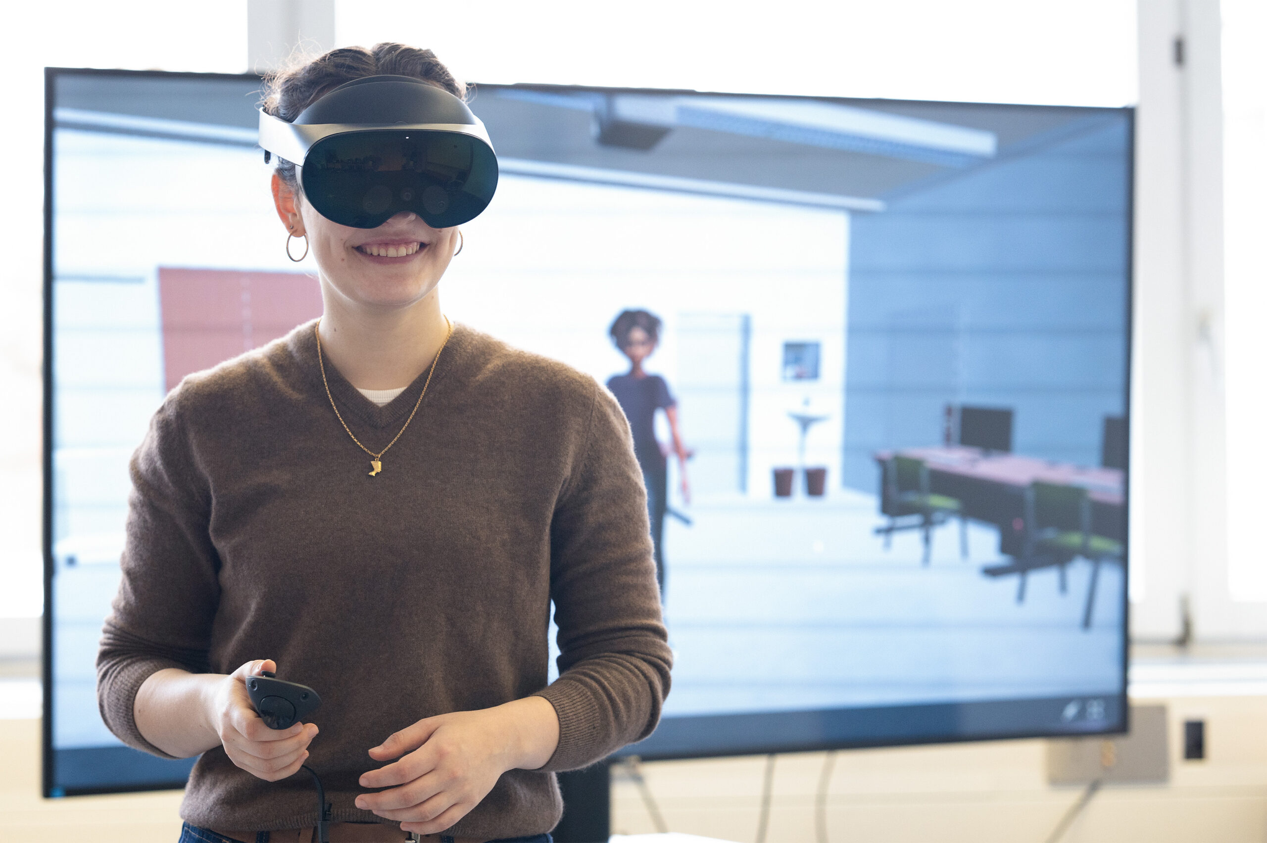 Eine Person, die ein VR-Headset trägt und einen Controller in der Hand hält, steht lächelnd vor einem Bildschirm, der einen virtuellen Raum mit einer digitalen Figur zeigt. at Hochschule Coburg