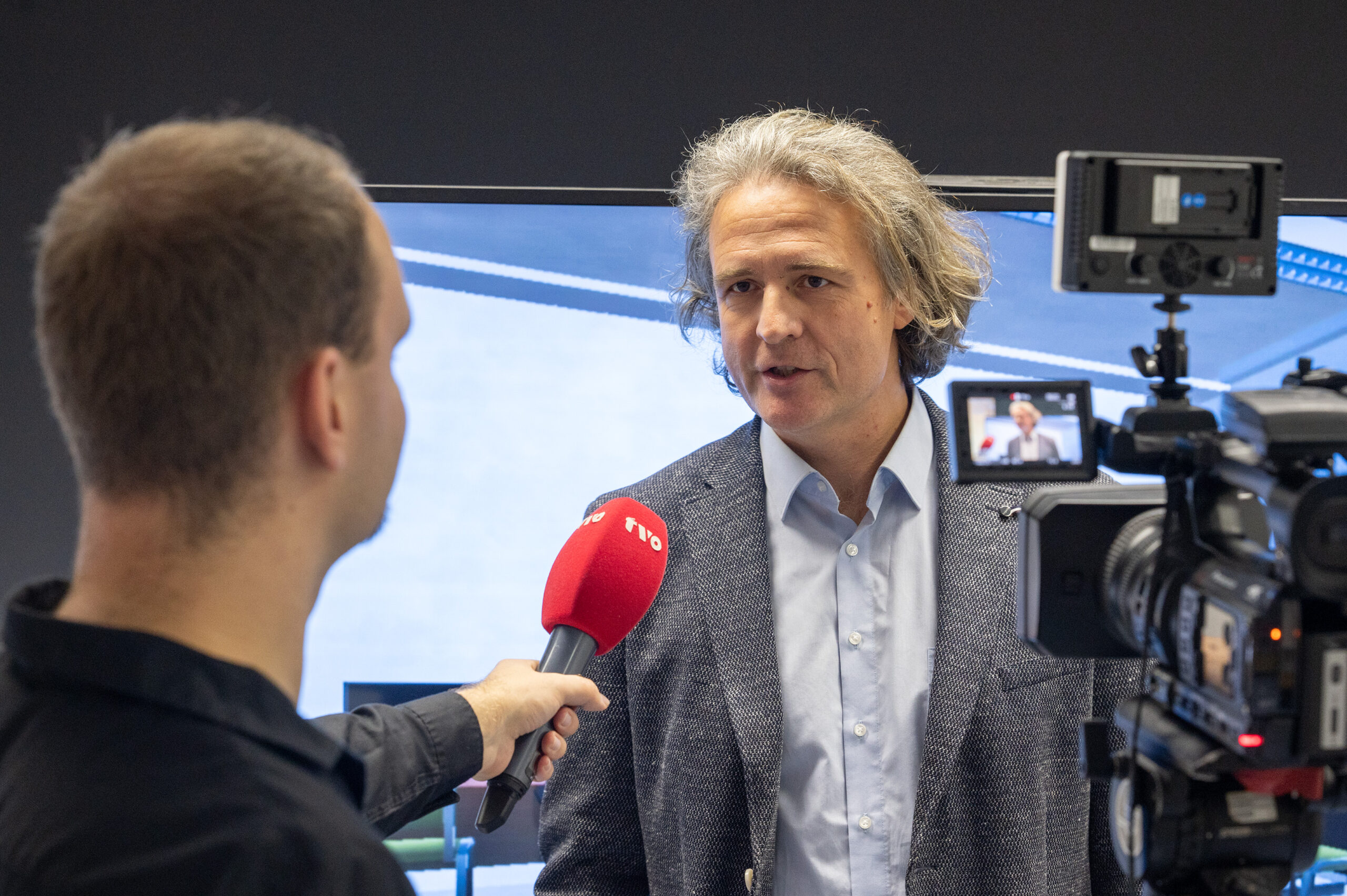 Ein Mann im Anzug wird von einem Reporter mit einem roten Mikrofon interviewt, während ein anderer für die Hochschule Coburg aufnimmt. In der professionellen Indoor-Kulisse werden Weiterentwicklungen wie der KI-Lerncoach und die virtuelle Tutorin vorgestellt. at Hochschule Coburg