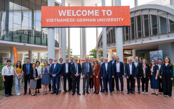 Eine Gruppe von Menschen posiert für ein Foto unter einem großen roten Banner mit der Aufschrift "WELCOME TO VIETNAMESE-GERMAN UNIVERSITY" vor einem modernen Universitätsgebäude in Ho-Chi-Minh, um die Partnerschaften mit den Bayerischen Hochschulen zu feiern. at Hochschule Coburg