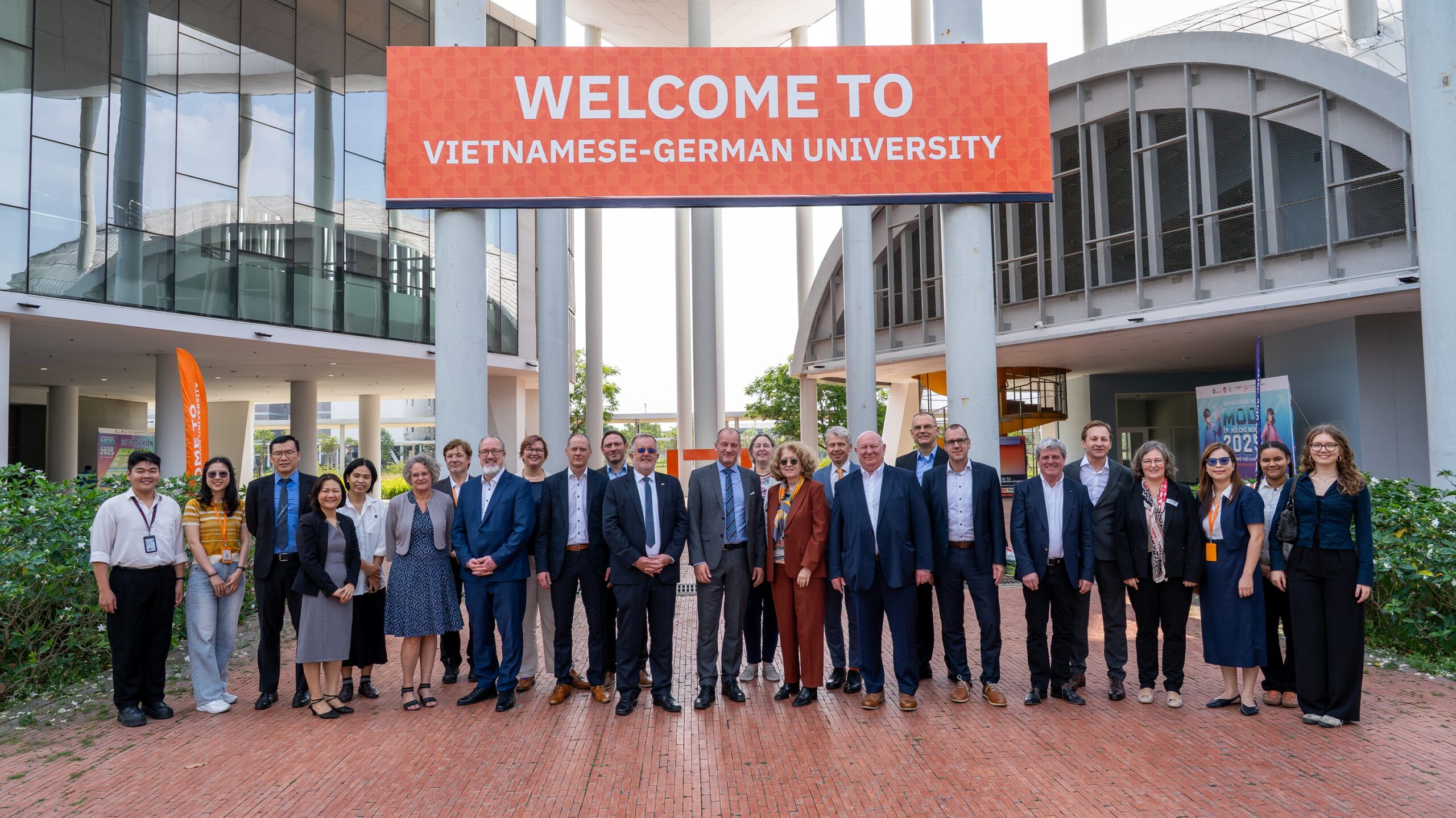 Eine Gruppe von Menschen posiert für ein Foto unter einem großen roten Banner mit der Aufschrift "WELCOME TO VIETNAMESE-GERMAN UNIVERSITY" vor einem modernen Universitätsgebäude in Ho-Chi-Minh, um die Partnerschaften mit den Bayerischen Hochschulen zu feiern. at Hochschule Coburg