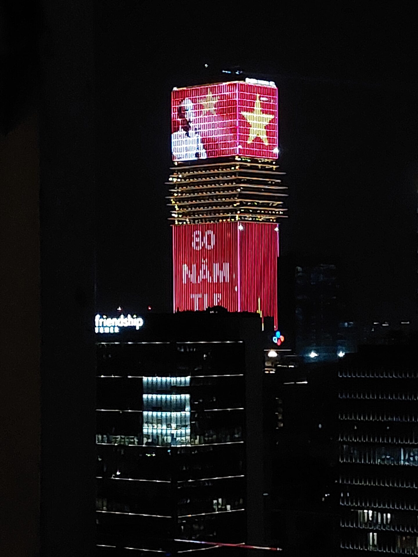 Ein hohes Gebäude bei Nacht zeigt rote und gelbe Lichter an seiner Fassade, darunter einen großen gelben Stern, ein Porträt von Ho-Chi-Minh und die Worte "HỘI QUÂN". Die umliegenden Gebäude sind dunkel mit einigen beleuchteten Fenstern, die sinnvolle Partnerschaften symbolisieren. at Hochschule Coburg