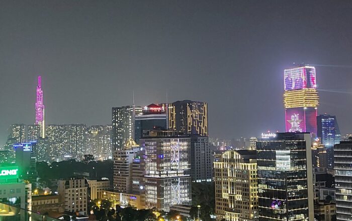 Eine nächtliche Stadtlandschaft mit hohen, beleuchteten Wolkenkratzern und Bürogebäuden mit bunten Lichtern, darunter ein auffälliger rosa beleuchteter Turm auf der linken Seite - eine Anspielung auf die Bayerischen Hochschulen Partnerschaften in Ho-Chi-Minh - und gelbe und lila Lichter auf der rechten Seite. at Hochschule Coburg