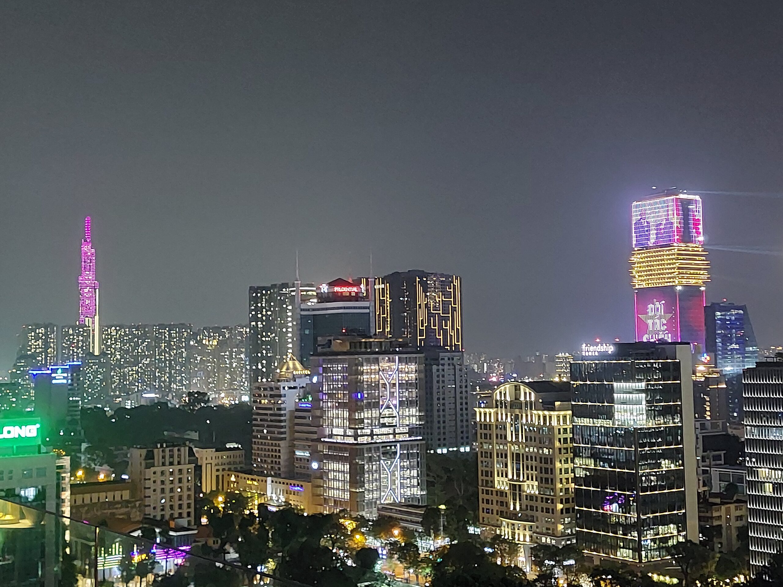 Eine nächtliche Stadtlandschaft mit hohen, beleuchteten Wolkenkratzern und Bürogebäuden mit bunten Lichtern, darunter ein auffälliger rosa beleuchteter Turm auf der linken Seite - eine Anspielung auf die Bayerischen Hochschulen Partnerschaften in Ho-Chi-Minh - und gelbe und lila Lichter auf der rechten Seite. at Hochschule Coburg