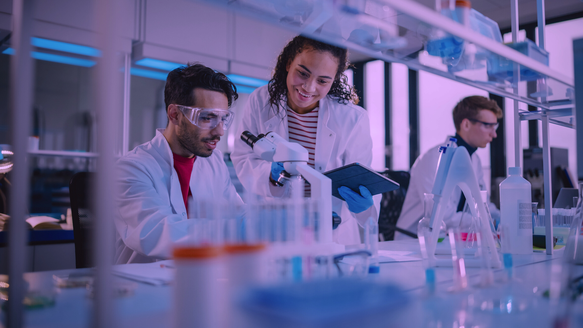 Zwei Wissenschaftler in Laborkitteln und mit Schutzbrillen arbeiten während der Langen Nacht der Wissenschaften an einem Labortisch. Der eine benutzt ein Mikroskop, während der andere ein Tablet hält. Ein weiterer Wissenschaftler arbeitet im Hintergrund inmitten verschiedener Laborgeräte. at Hochschule Coburg