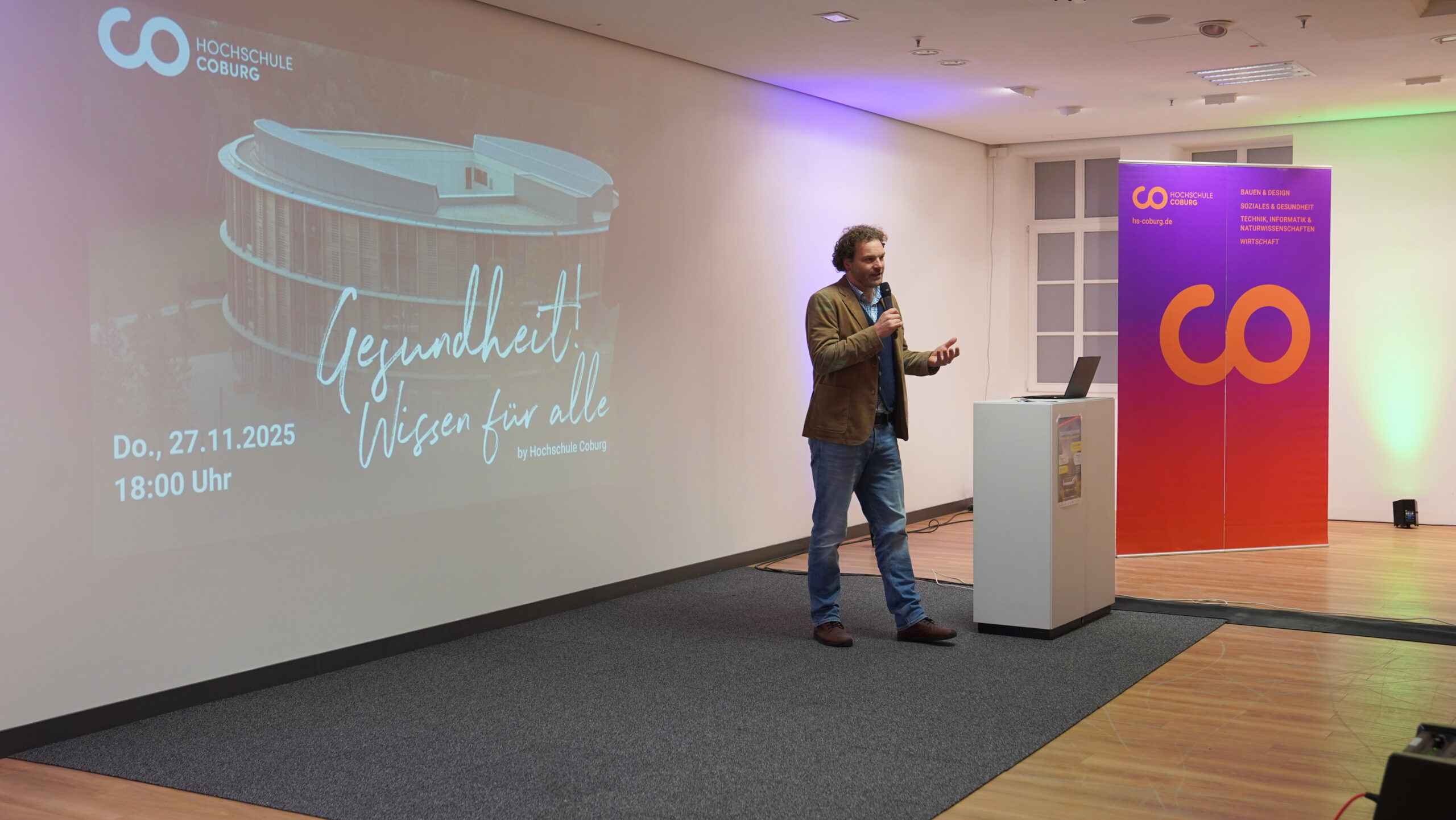 Ein Mann in einer braunen Jacke spricht auf der Bühne in ein Mikrofon. Hinter ihm zeigt ein großer Bildschirm die Veranstaltungshinweise zum Krankenhausalltag an der Hochschule Coburg am 27. November 2025. An der Seite stehen leuchtende Transparente. at Hochschule Coburg