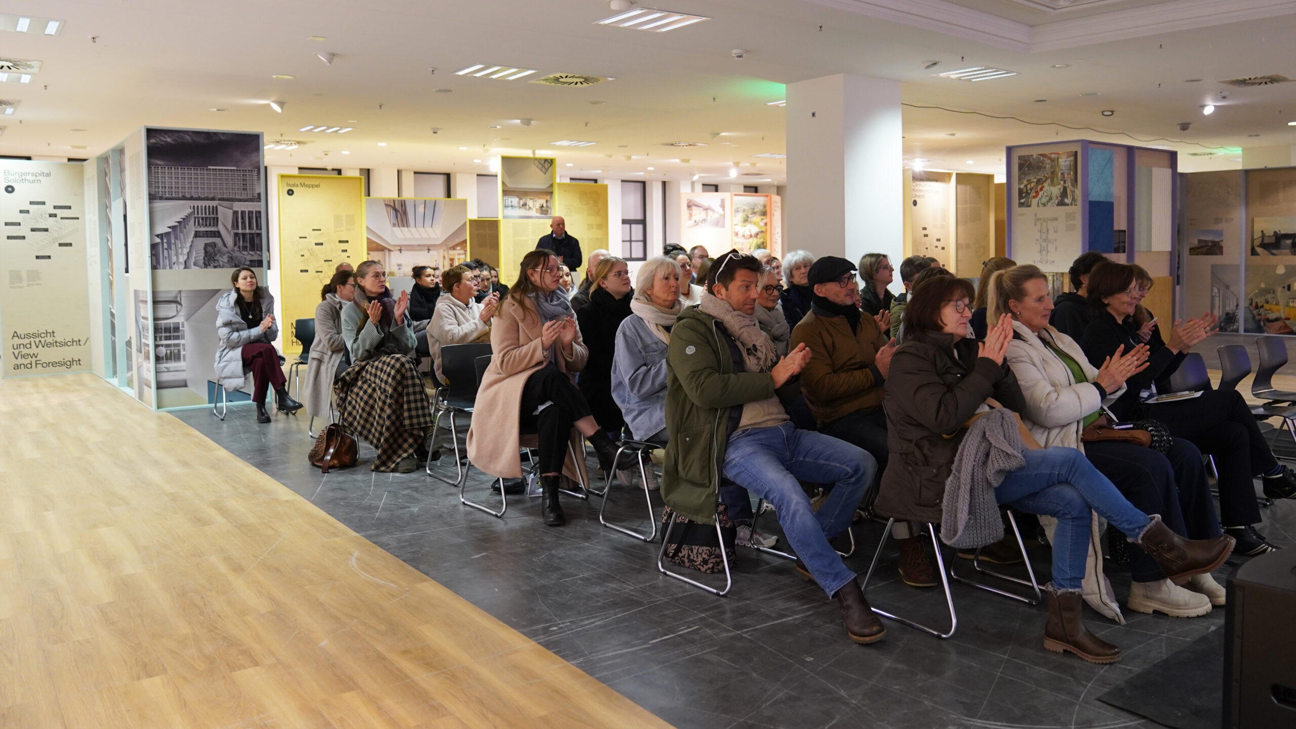 Eine Gruppe von Menschen sitzt auf Stühlen in einem gut beleuchteten Ausstellungsraum, aufmerksam nach vorne blickend; einige klatschen, während andere zuhören oder zuschauen, während im Hintergrund Schautafeln und Fotos zum Krankenhausalltag und zur heilenden Architektur zu sehen sind. at Hochschule Coburg