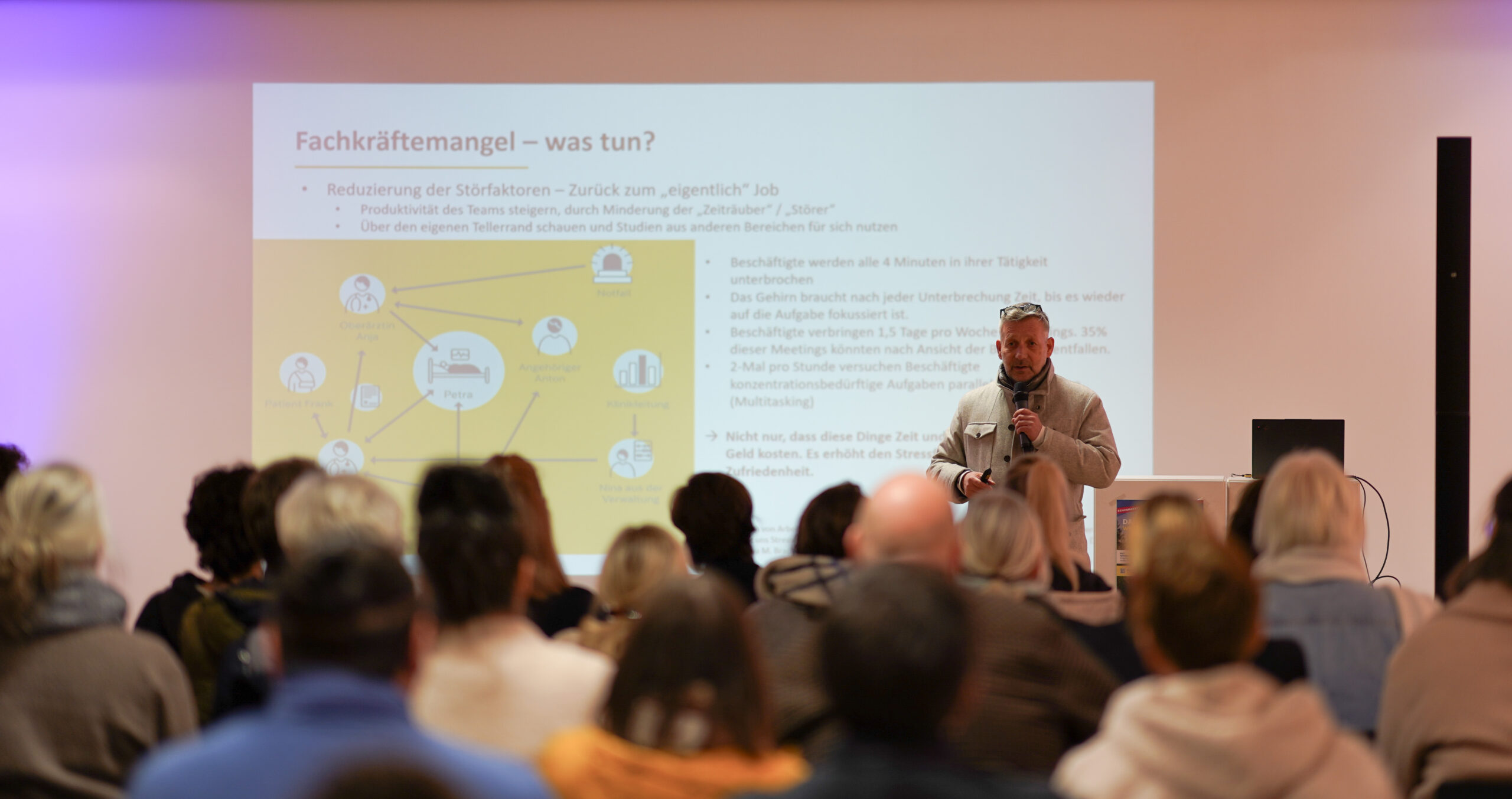 Ein Redner steht vor einem sitzenden Publikum und präsentiert eine Folie auf Deutsch über Arbeitskräftemangel und Lösungen im Krankenhausalltag. Die Folie zeigt einen Titel, Aufzählungspunkte und eine Infografik mit verbundenen Symbolen. at Hochschule Coburg