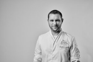 Ein Mann in einer Kochjacke steht vor einem schlichten Hintergrund, blinzelt mit einem Auge und lächelt leicht. Auf der Jacke steht "Daniel Klein" und "Oskar Hacker Stiftung". Das schwarz-weiße Bild fängt seine Selbstmotivation ein. at Hochschule Coburg