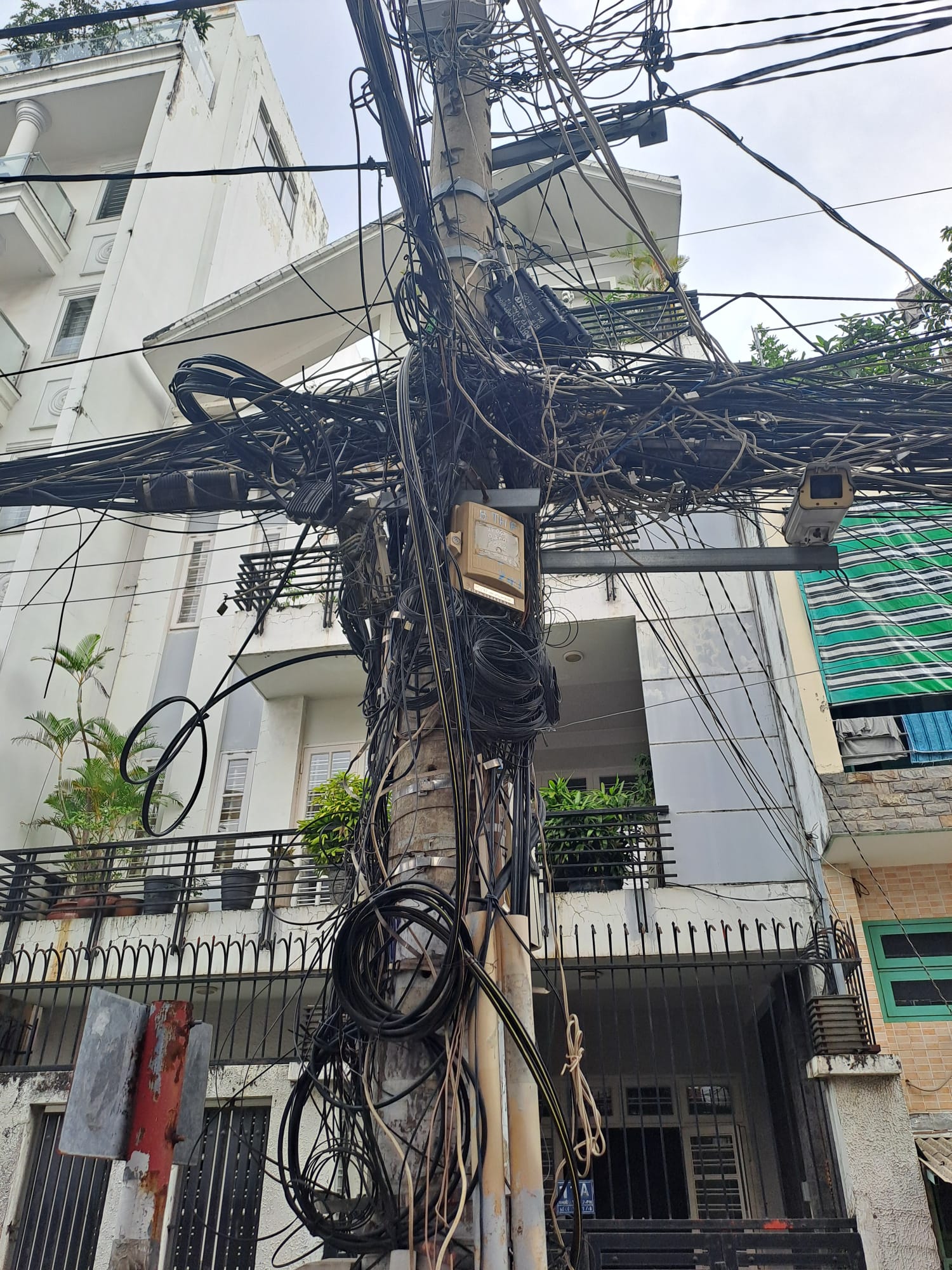 Ein Strommast mit einem Wirrwarr aus elektrischen Leitungen und Kabeln steht chaotisch vor einem Gebäude in Ho-Chi-Minh. Die vielen hängenden Drähte spiegeln die Komplexität der städtischen Infrastruktur und der globalen Partnerschaften wider. at Hochschule Coburg