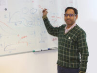 A man wearing glasses and a green checkered sweater is standing by a whiteboard, holding a marker and smiling. The whiteboard is filled with diagrams, graphs, and handwritten notes. at Hochschule Coburg