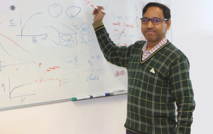 A man wearing glasses and a green checkered sweater is standing by a whiteboard, holding a marker and smiling. The whiteboard is filled with diagrams, graphs, and handwritten notes. at Hochschule Coburg