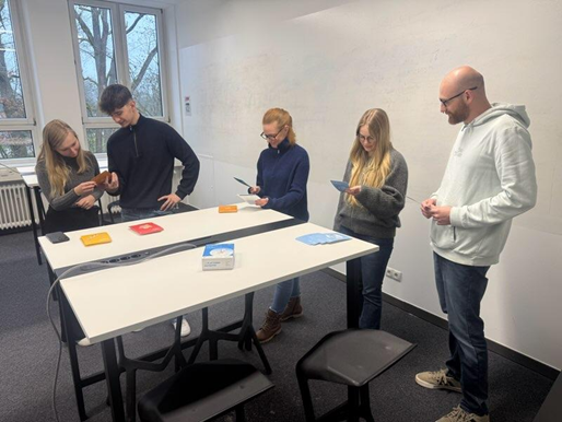 Fünf Personen stehen in einem hellen Raum um einen hohen Tisch herum, jeder hält eine Karte in der Hand und ist mit einer vom Studium inspirierten Tätigkeit beschäftigt. Spielschachteln und Karten liegen verstreut auf dem Tisch, während große Fenster im Hintergrund für natürliches Licht sorgen. at Hochschule Coburg