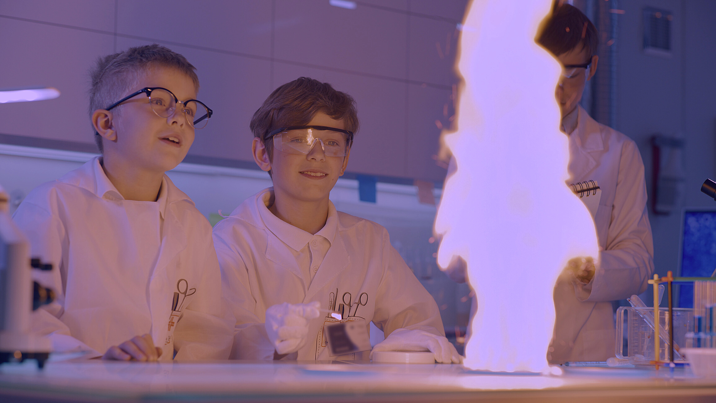 Zwei Kinder in Laborkitteln und mit Schutzbrillen beobachten eine große blaue Flamme auf einem Labortisch. Sie wirken aufgeregt und beschäftigen sich mit einem wissenschaftlichen Experiment während der Langen Nacht der Wissenschaften. Im Hintergrund ist eine Laborausrüstung zu sehen. at Hochschule Coburg