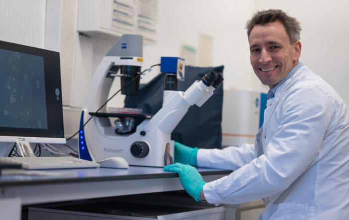 Ein lächelnder Wissenschaftler in einem weißen Laborkittel und mit blaugrünen Handschuhen sitzt an einem Labortisch und benutzt ein Mikroskop. Auf einem Computermonitor neben ihm werden Zellbilder aus der aktuellen Forschung zur Biologie der Verjüngung angezeigt. Die Laborumgebung wirkt sauber und aufgeräumt. at Hochschule Coburg
