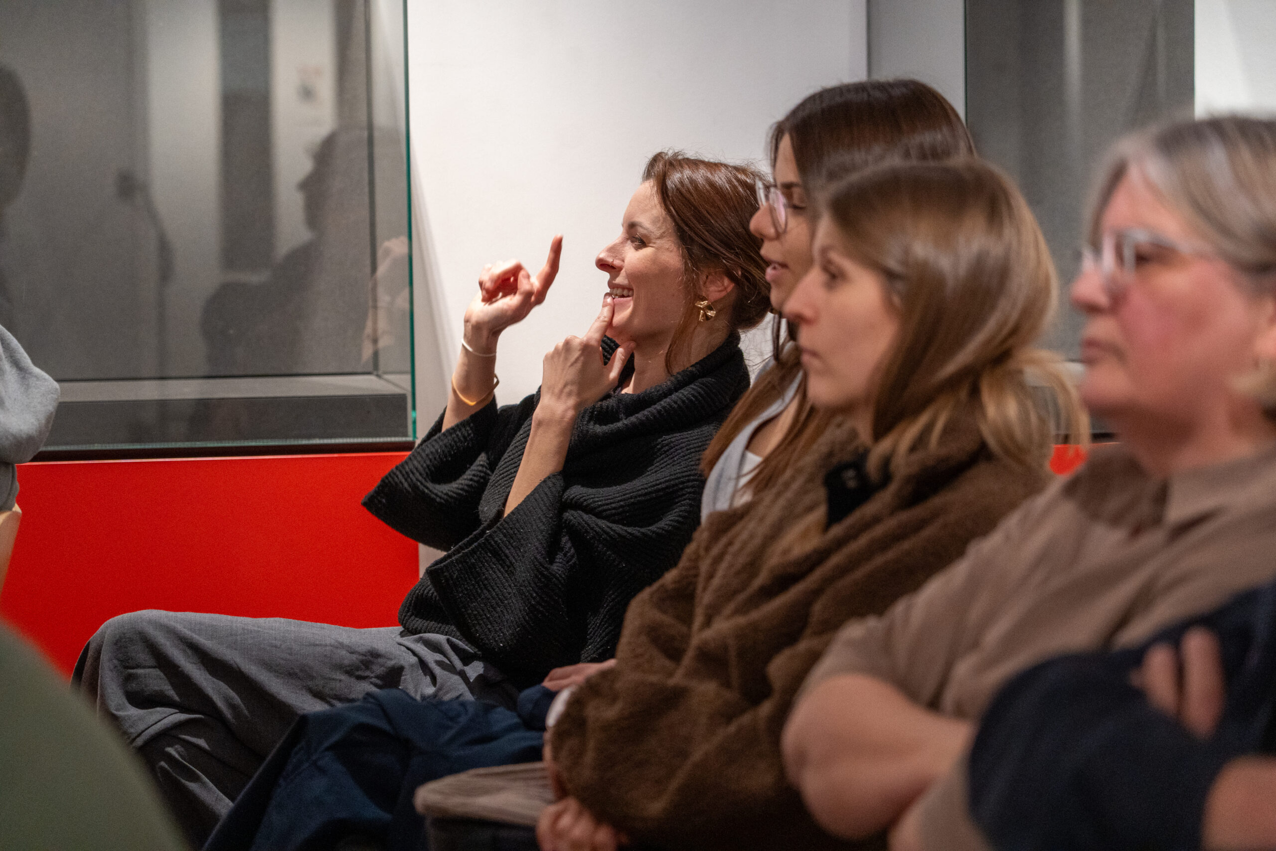 Eine Gruppe von Frauen sitzt in einem Raum und hört aufmerksam zu; eine Frau in der Mitte lächelt und gestikuliert mit den Händen, möglicherweise in Gebärdensprache. Die Atmosphäre scheint konzentriert zu sein, während sie über Entwürfe und mögliche Zukünfte diskutieren. at Hochschule Coburg