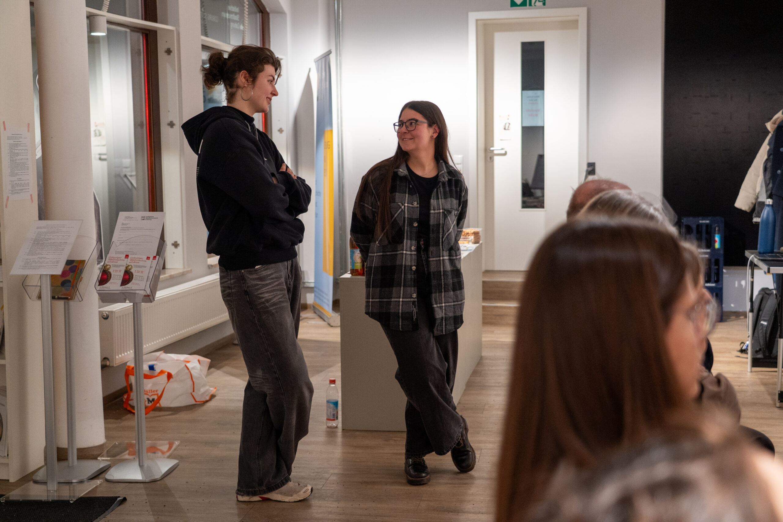Zwei Frauen stehen in einem hell erleuchteten Innenraum und lächeln sich an. Lässig gekleidet, teilen sie einen entspannten Moment, während die Menschen im Vordergrund über Entwürfe und Zukünfte plaudern und eine warme und soziale Atmosphäre schaffen. at Hochschule Coburg