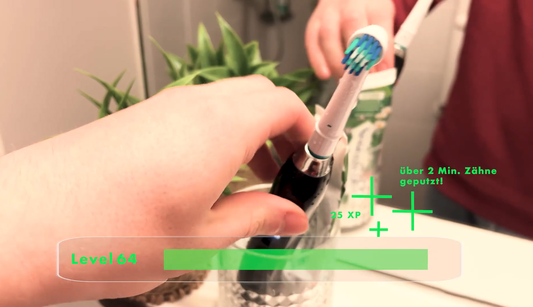 Eine Hand stellt eine elektrische Zahnbürste in ein Glas, während grüne Grafiken das Bild überlagern und auf mögliche Zukünfte des spielerischen Zähneputzens hinweisen. "Level 64", "25 XP" und "über 2 Min. Zähne geputzt!" erscheinen, mit einer Pflanze und einem Waschbecken im Hintergrund. at Hochschule Coburg