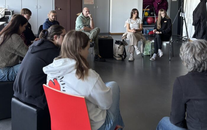 Eine Gruppe von Menschen sitzt in einem Kreis in einem modernen Raum und führt eine konzentrierte Diskussion. Die meisten sind leger gekleidet, einige haben Notebooks oder Taschen dabei, was die Menschlichkeit und Offenheit einer demokratischen Gesellschaft widerspiegelt. at Hochschule Coburg