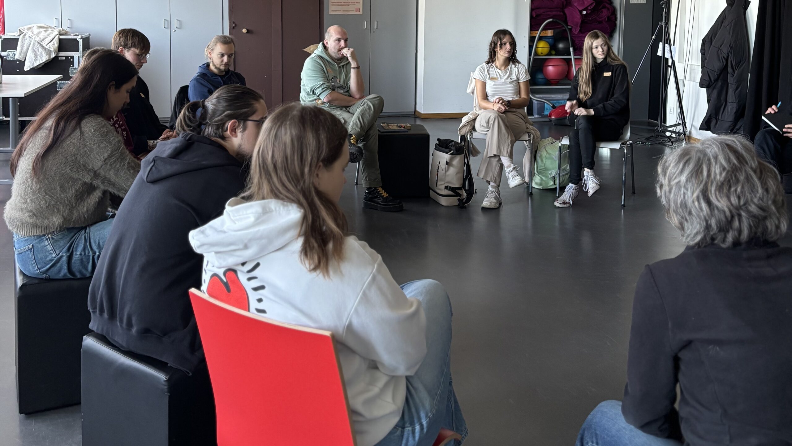 Eine Gruppe von Menschen sitzt in einem Kreis in einem modernen Raum und führt eine konzentrierte Diskussion. Die meisten sind leger gekleidet, einige haben Notebooks oder Taschen dabei, was die Menschlichkeit und Offenheit einer demokratischen Gesellschaft widerspiegelt. at Hochschule Coburg