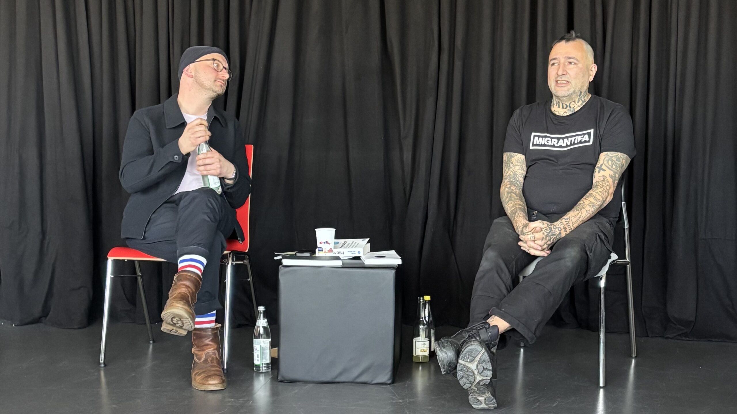 Zwei Männer sitzen auf Stühlen vor einem schwarzen Vorhang. Der eine, mit Hut und bunten Socken, und der andere, tätowiert mit einem "Migranti"-Shirt, teilen sich Getränke und Papiere - ein Spiegelbild der Menschlichkeit in einer demokratischen Gesellschaft. at Hochschule Coburg