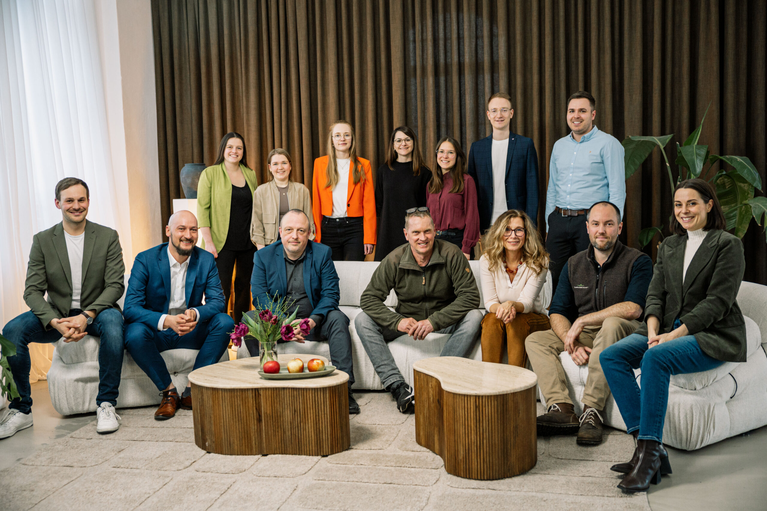 Eine Gruppe von fünfzehn Personen posiert in einem modernen Wohnzimmer, versammelt um hölzerne Couchtische mit Obst und Blumen - vielleicht diskutieren sie über Forschung für Kommunen - vor braunen Vorhängen und üppigen Zimmerpflanzen. at Hochschule Coburg