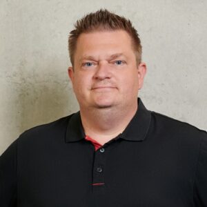 A man with short brown hair wearing a black polo shirt stands in front of a plain, light-colored wall, looking at the camera with a neutral expression. at Hochschule Coburg