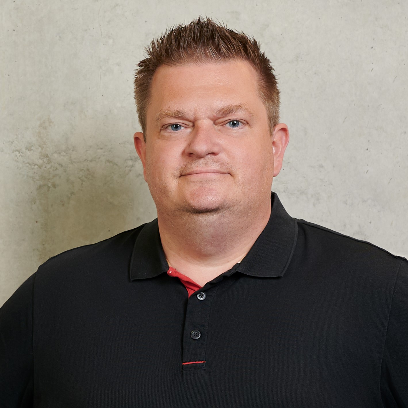 Ein Mann mit kurzen, hellbraunen Haaren und einem schwarzen Polohemd steht vor einer schlichten, hellen Wand und blickt mit neutralem Blick in die Kamera. at Hochschule Coburg