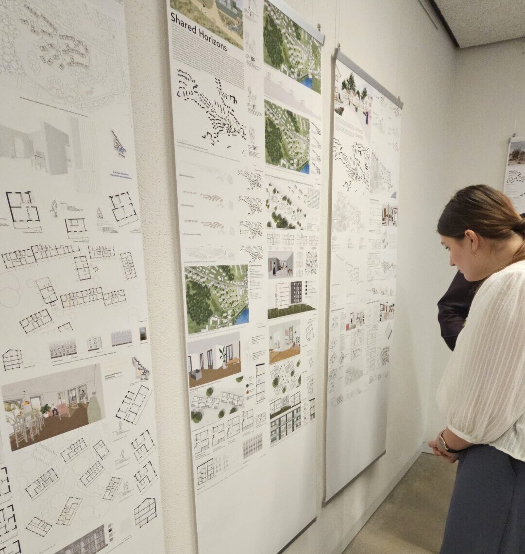 Eine Person betrachtet aufmerksam die an einer weißen Wand angebrachten architektonischen Entwurfstafeln mit Grundrissen, Bildern und Diagrammen in einer Ausstellung, die neue Perspektiven auf die Stadtentwicklung der Nachkriegszeit bietet. at Hochschule Coburg