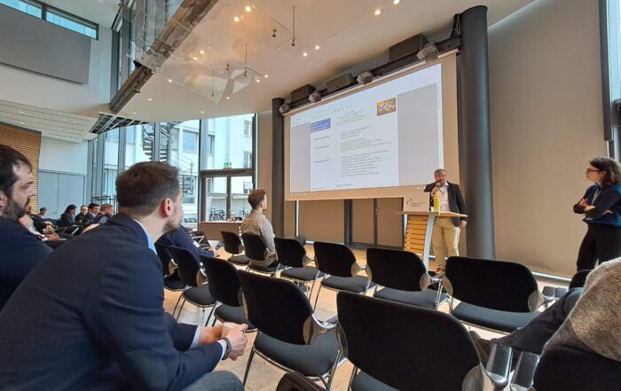 Ein Mann steht an einem Podium und präsentiert einem Publikum, das in einem modernen Konferenzraum mit großen Fenstern und schwarzen Stuhlreihen sitzt, Folien über europäische Forschungsprojekte und pflegt neue Kontakte unter den Teilnehmern. at Hochschule Coburg