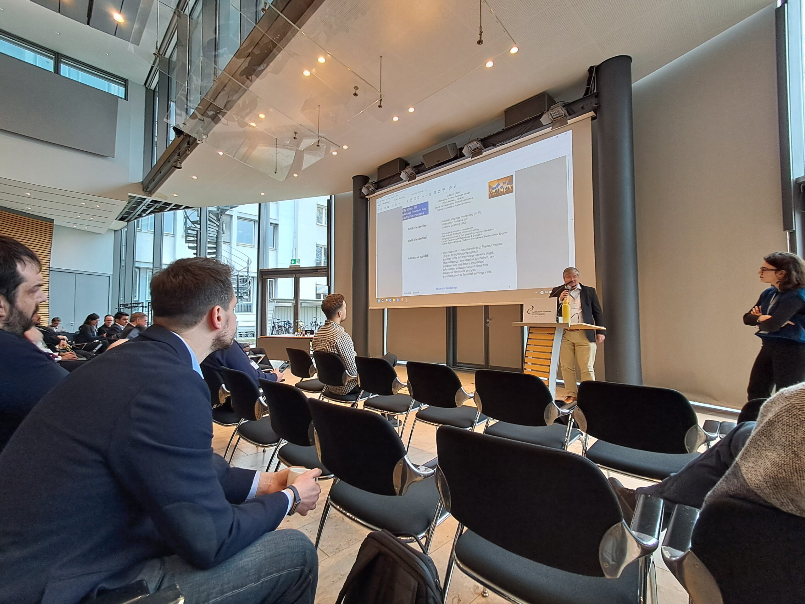 Ein Mann steht an einem Podium und präsentiert einem Publikum, das in einem modernen Konferenzraum mit großen Fenstern und schwarzen Stuhlreihen sitzt, Folien über europäische Forschungsprojekte und pflegt neue Kontakte unter den Teilnehmern. at Hochschule Coburg