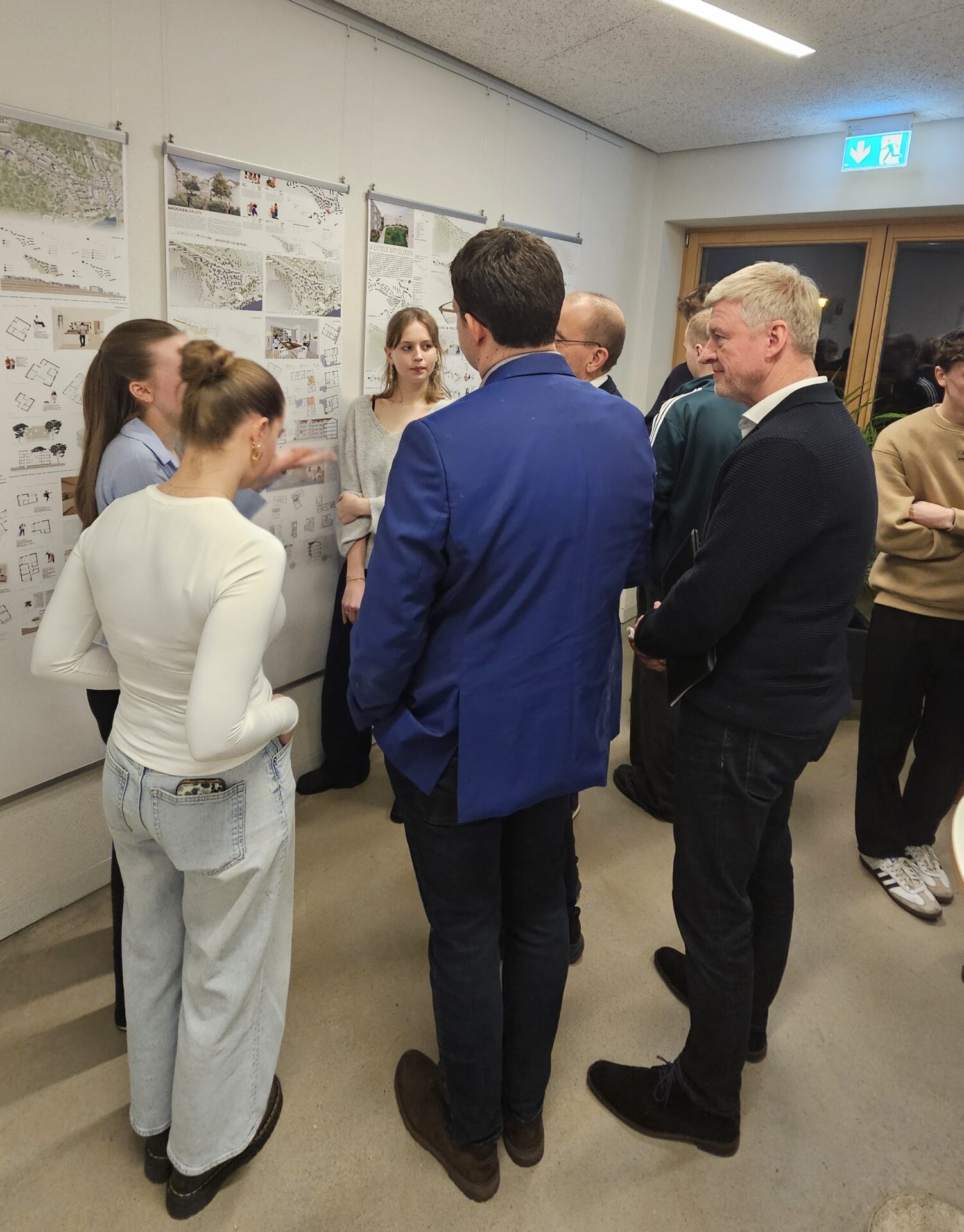 Eine Gruppe von Menschen steht vor einer Wand mit Architekturzeichnungen und Karten in der Ausstellung und diskutiert über neue Perspektiven. Einige stehen mit dem Gesicht zur Wand, während andere zuhören und sich unterhalten. at Hochschule Coburg